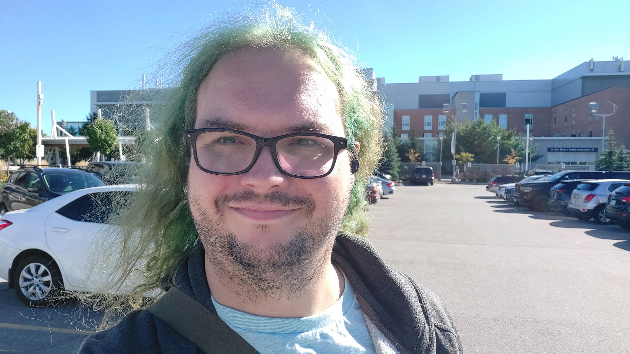 A man with long green hair stands in a hospital parking lot, hospital in the background, with a clear blue sky